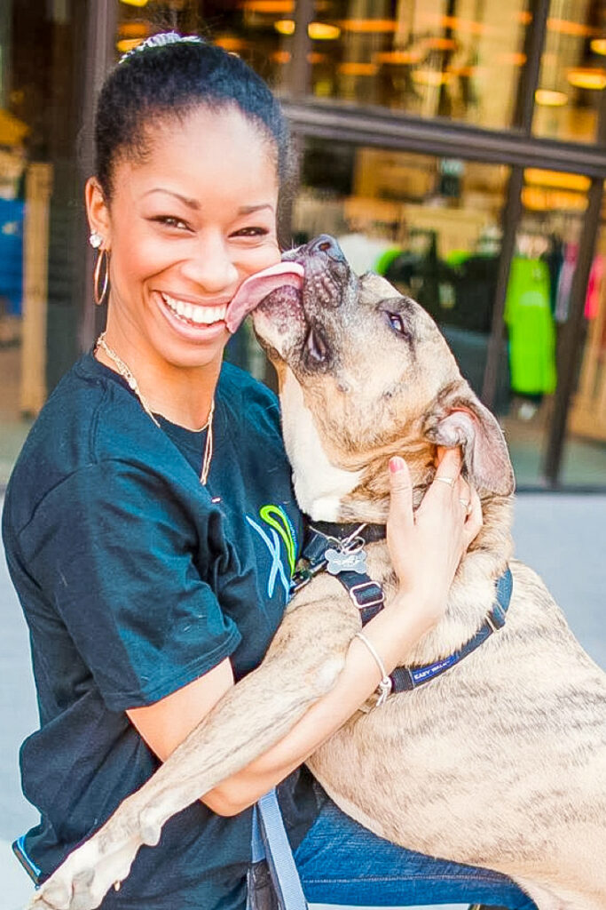 Victoria Vaden, a dog mom, smiling with a pit bull boxer mix dog, in front of a store as he licks her cheek